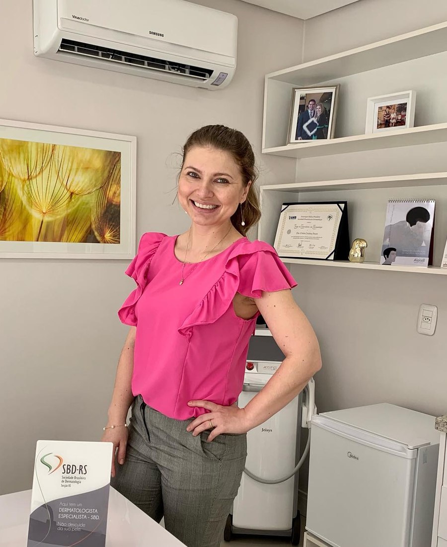 Dra. Cíntia Pessin em pé no consultório, sorrindo, com equipamento de laser ao fundo