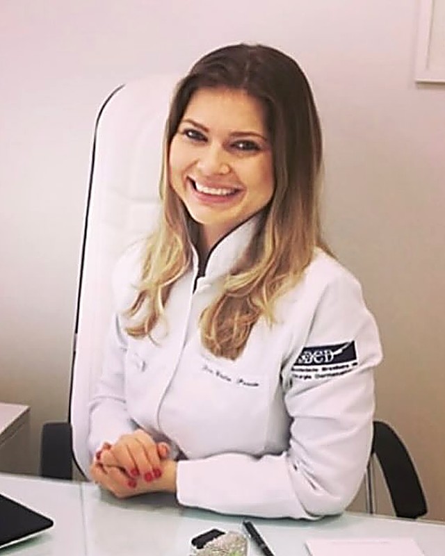 Dra. Cíntia Pessin de jaleco, sentada à mesa do consultório, sorrindo para a câmera